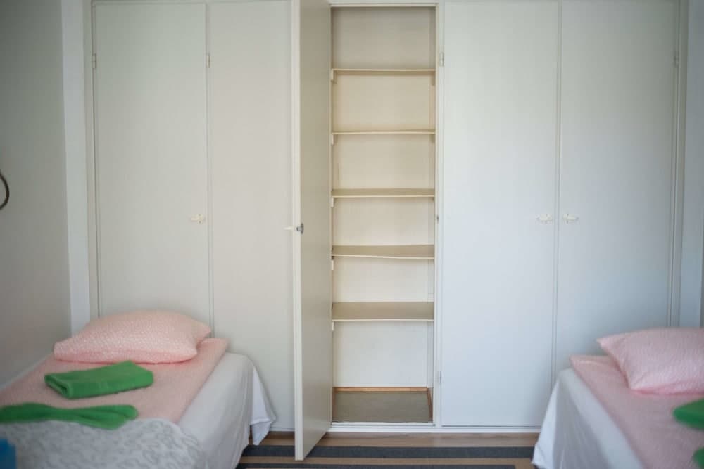 A cozy dormitory room features twin beds with pink bedding and green towels, a large open closet with empty shelves, and a minimalist design ideal for budget travelers seeking simple accommodations.
