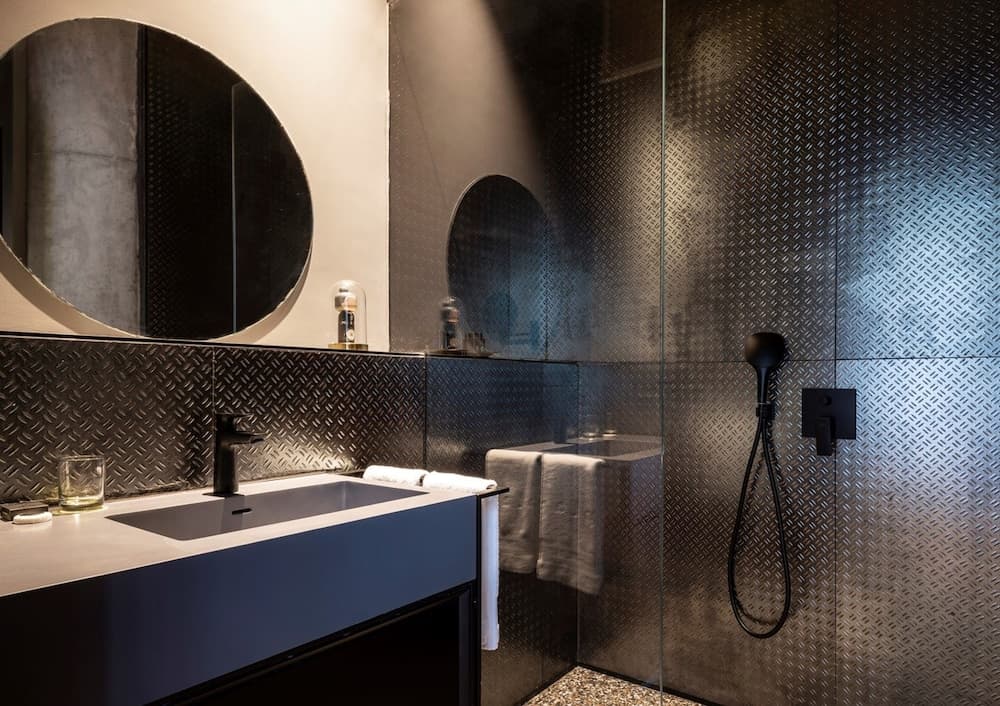 A modern bathroom with sleek metallic walls, a minimalist vanity, and a round mirror exudes an industrial chic vibe, perfect for travelers seeking a stylish accommodation experience.