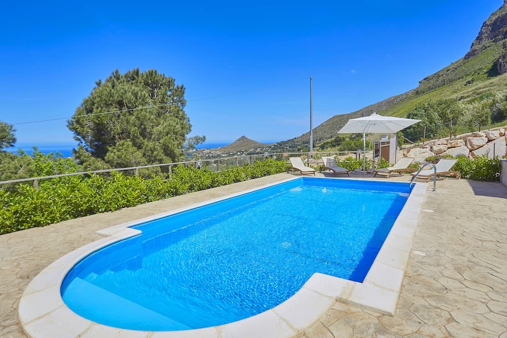 A serene private pool area with lounge chairs and an umbrella overlooks lush greenery and distant hills under a clear blue sky, ideal for a relaxing vacation retreat.