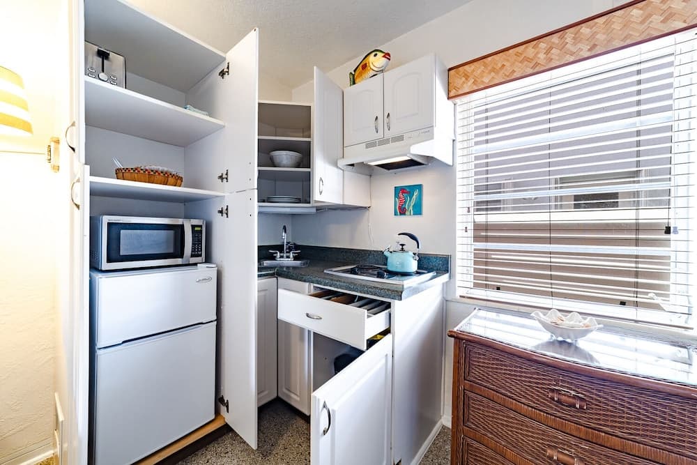A cozy, compact kitchenette with white cabinetry, a microwave, and a teal kettle on the stove, bathed in soft natural light from a window with woven blinds, ideal for travelers seeking a quaint and functional space for self-catering stays.