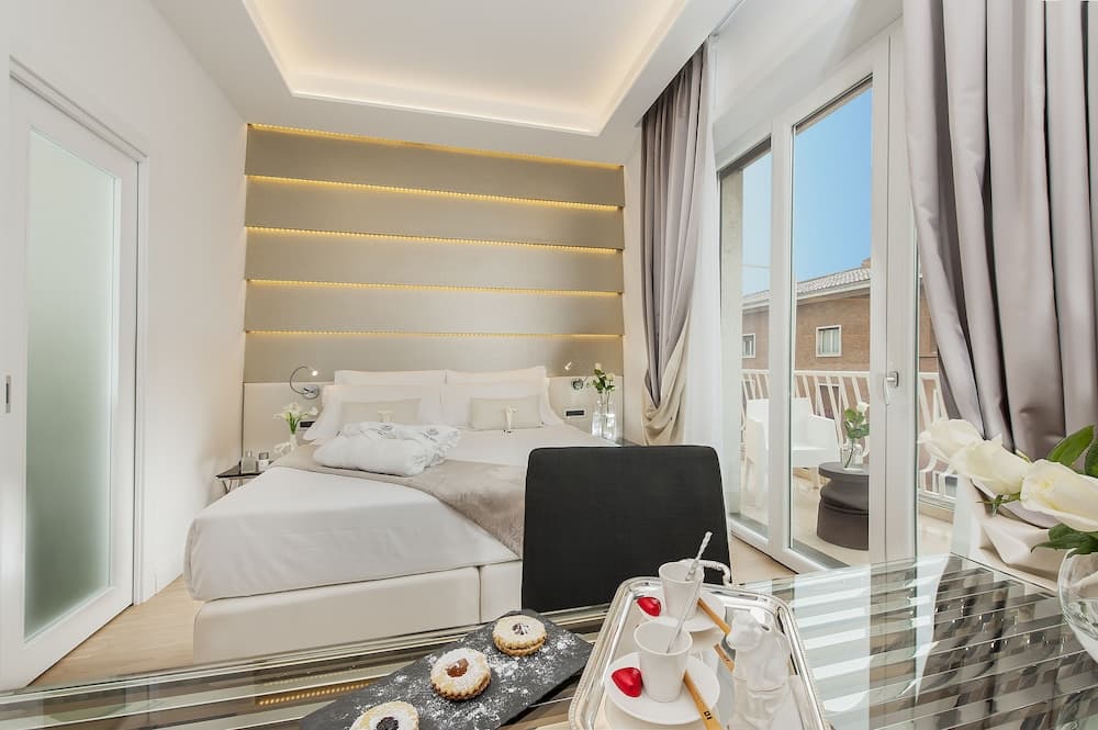 A serene hotel room features a cozy bed with a modern lighted headboard, a glass table with pastries and tea, and a balcony offering a bright outdoor view, ideal for travelers seeking relaxation and comfort.
