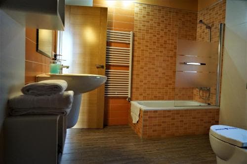 A cozy hotel bathroom features warm-toned tiled walls, a modern bathtub with a glass shower screen, neatly folded towels on a stool, and a sleek sink, ideal for travelers seeking comfort and style during their stay.