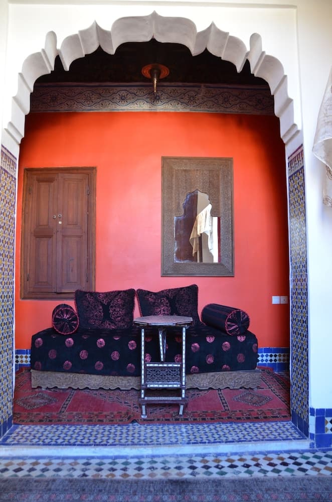 A vibrant orange-walled alcove adorned with intricate Moroccan tilework, plush black polka-dot cushions, and a carved wooden table creates an inviting and exotic atmosphere perfect for cultural exploration and relaxation.