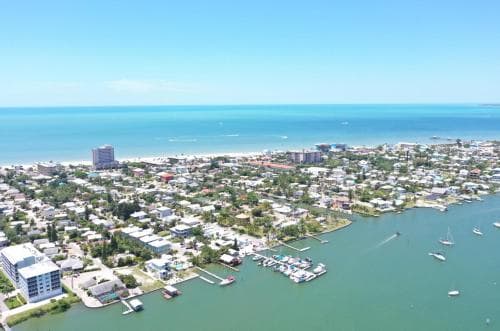 An aerial view of a vibrant coastal town with clear turquoise waters, docked boats, and dense residential areas under a sunny sky, ideal for beachside relaxation and waterfront exploration.