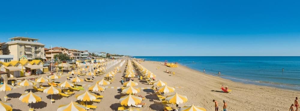 A vibrant beach scene with rows of yellow-and-white umbrellas and loungers stretches along the sandy shore under a clear blue sky, ideal for a relaxing seaside vacation.