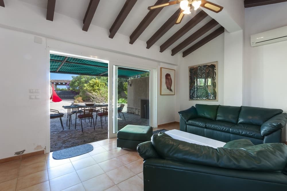 A cozy living room with green leather sofas and wooden beams opens to a serene outdoor patio with dining furniture, ideal for vacation rental seekers looking for relaxation and comfort.