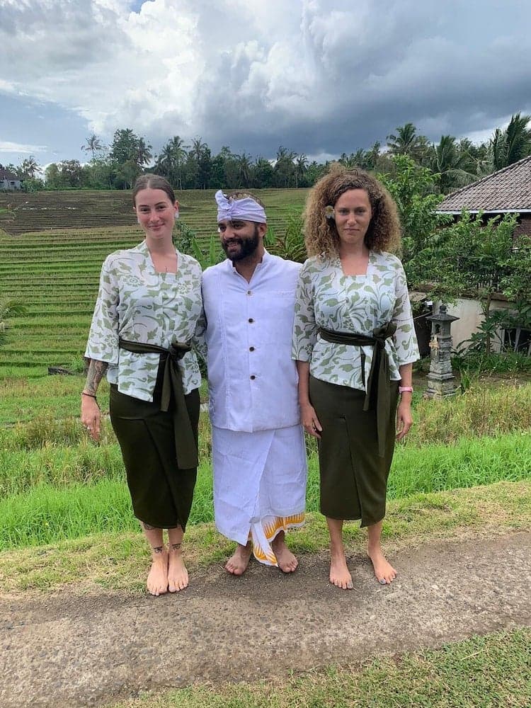 Three individuals dressed in traditional attire stand barefoot on a path near lush rice terraces under a cloudy sky, capturing a serene cultural experience ideal for travelers exploring rural landscapes.