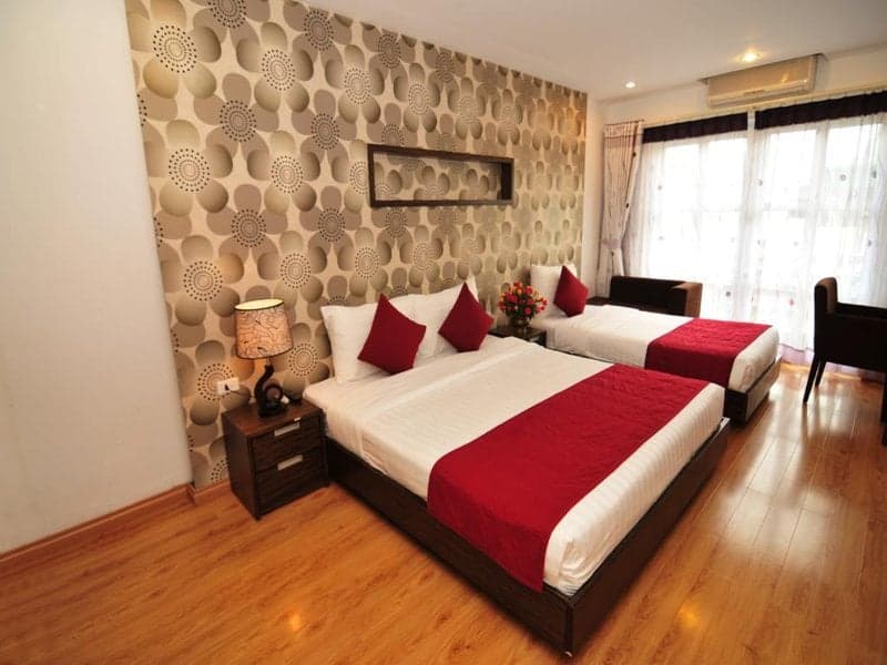 A cozy hotel room features two neatly made beds with red accents against a patterned wall, large windows letting in soft light, and wooden flooring, ideal for travelers seeking comfort and style.