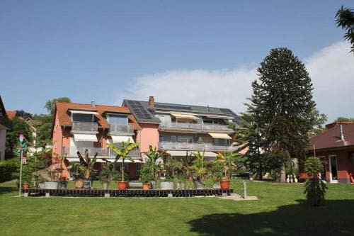 A serene suburban neighborhood features a charming apartment building adorned with balconies and solar panels, surrounded by lush greenery and potted plants, ideal for travelers seeking a peaceful and eco-friendly accommodation.