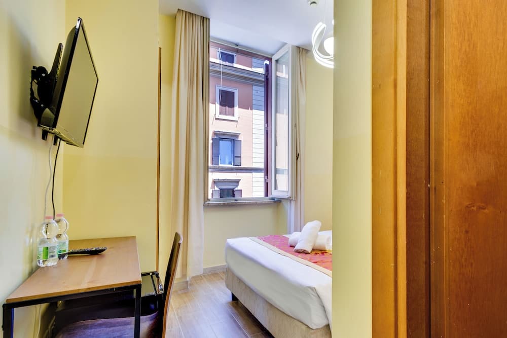 A cozy hotel room with yellow walls, a neatly made bed by the window, a desk with a mounted TV, and a serene urban view, ideal for travelers seeking comfort in a city stay.