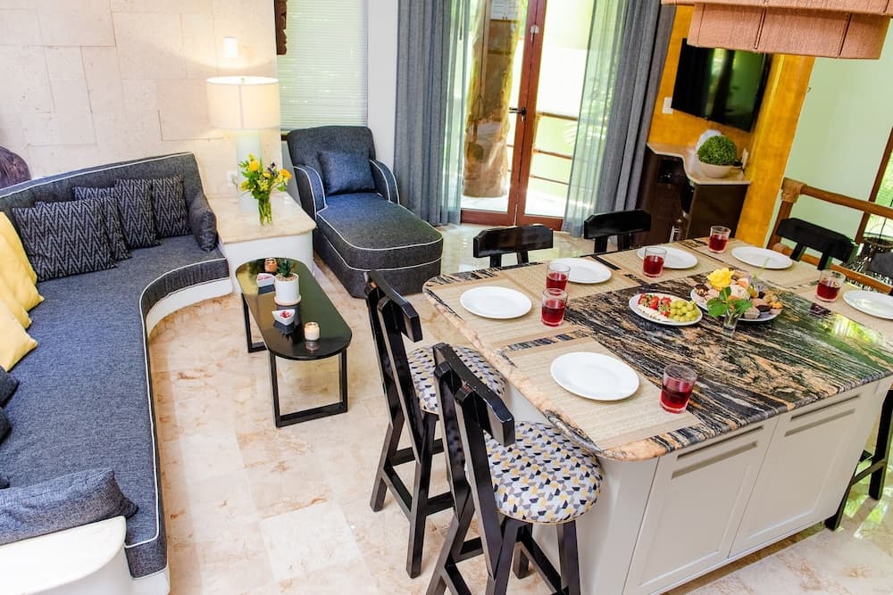 A cozy, well-lit living room and dining area with modern furnishings, vibrant decor, and a view of greenery outside through glass doors, ideal for travelers seeking a comfortable and inviting vacation rental space.