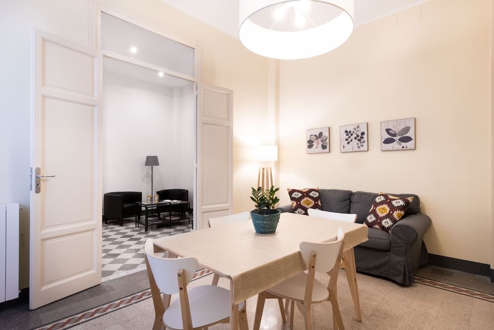 A cozy apartment living space features a modern dining table with white chairs, a gray sofa adorned with patterned cushions, and a checkered floor in the adjacent room, ideal for travelers seeking a comfortable urban retreat.