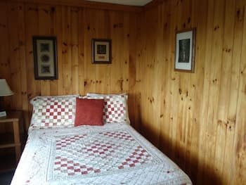 A cozy cabin bedroom with wooden walls, a checkered quilted bedspread, framed artwork, and warm lighting creates an inviting retreat perfect for a relaxing getaway.
