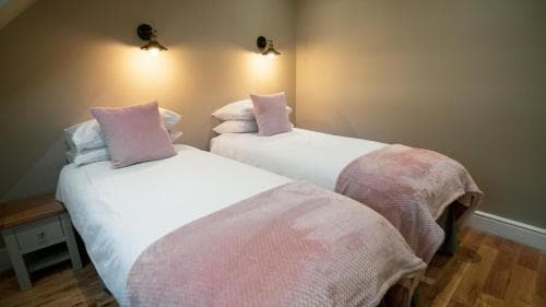 A cozy guestroom with two neatly arranged twin beds adorned with pink throws and pillows, illuminated by warm wall sconces, creates an inviting atmosphere ideal for travelers seeking comfort in a boutique accommodation setting.