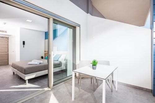 A modern, minimalist apartment features a sleek dining area with a small table and chairs, adjacent to a cozy bedroom visible through glass sliding doors, ideal for travelers seeking stylish urban accommodations.