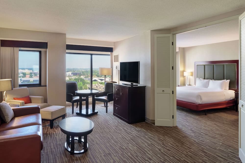 A cozy hotel suite features a spacious living area with leather seating, a flat-screen TV, and a bedroom with a neatly made bed, all overlooking a scenic cityscape through large windows, ideal for travelers seeking comfort and relaxation.