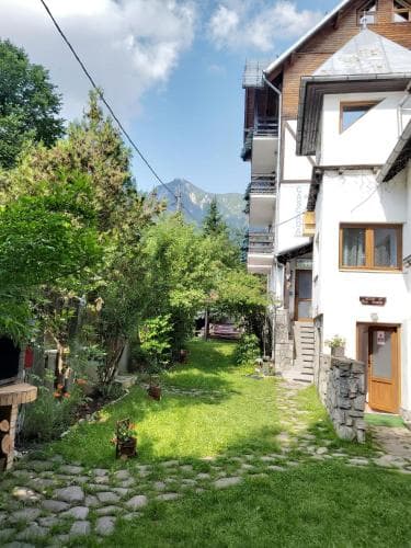 A serene courtyard garden with lush greenery and stone pathways invites travelers to relax near quaint white houses with balconies, set against the backdrop of majestic mountains, ideal for a peaceful getaway.