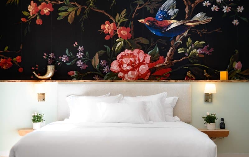 A cozy hotel room features a bed adorned with crisp white linens beneath a vibrant floral mural with a blue bird, creating an inviting atmosphere perfect for travelers seeking comfort and artistic ambiance.