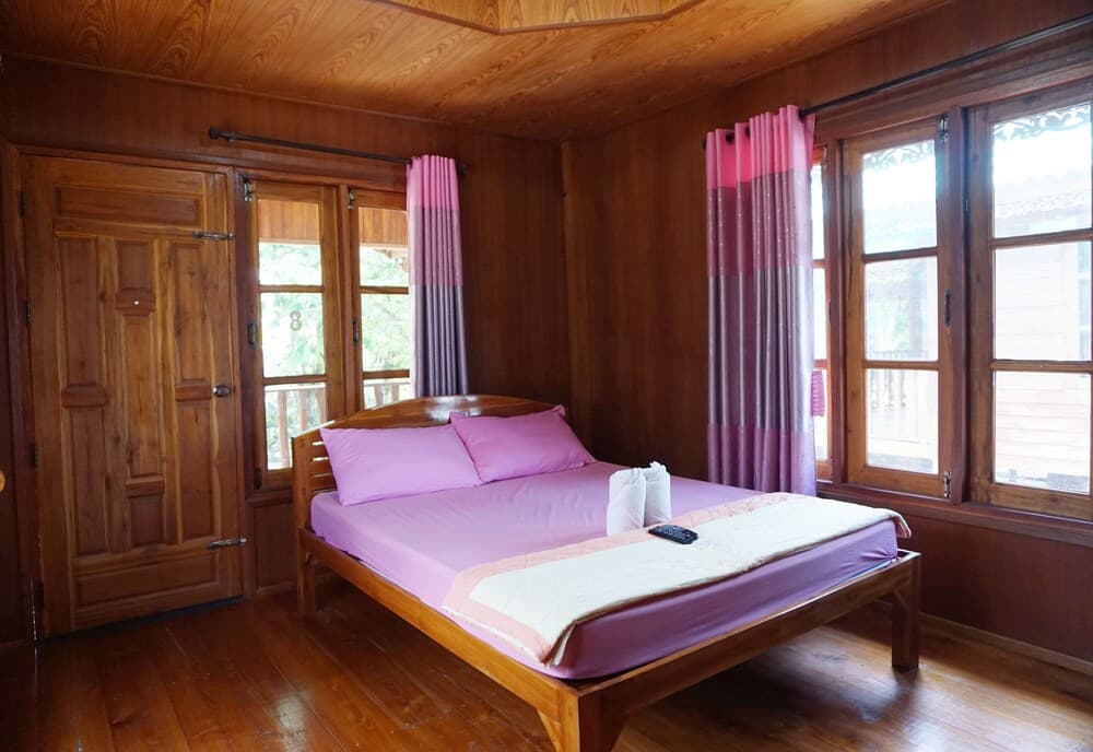 A cozy wooden cabin bedroom with vibrant purple bedding and curtains, featuring large windows and a serene outdoor view, ideal for travelers seeking a tranquil retreat.