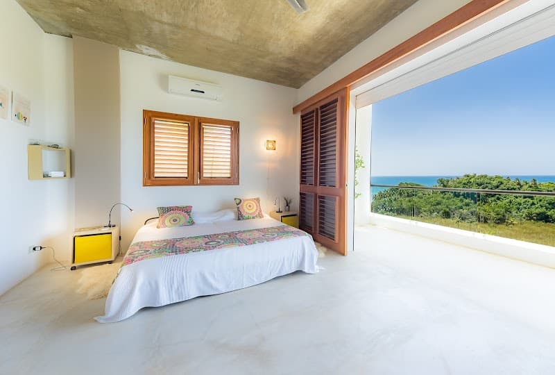 A serene bedroom with vibrant bedding overlooks lush greenery and the ocean from a spacious balcony, ideal for a tranquil retreat in Las Terrenas, Dominican Republic.