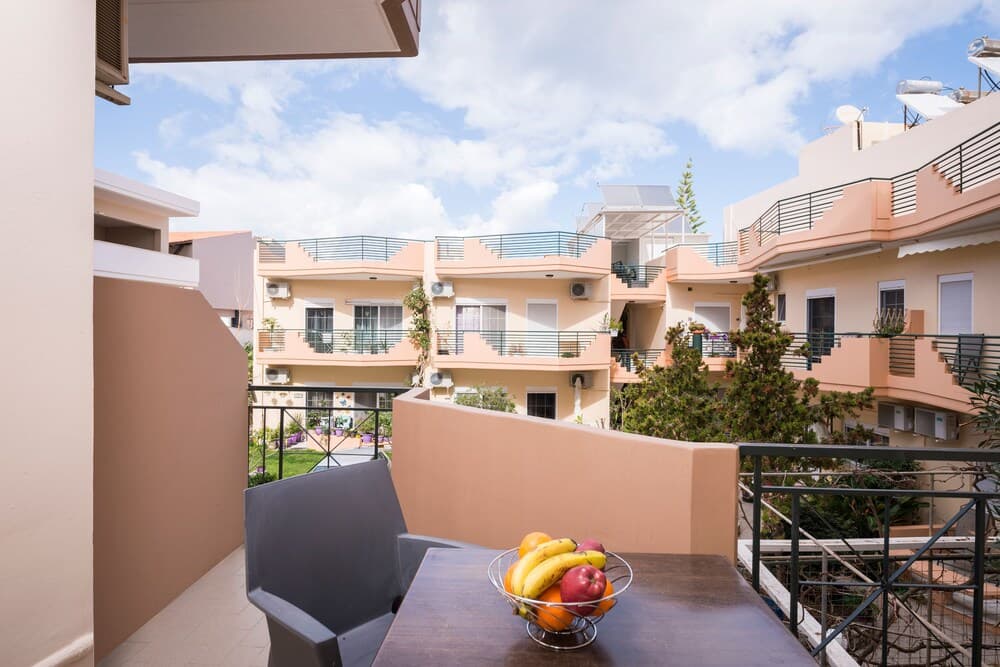 A serene balcony scene with a fruit bowl on a table, overlooking pastel-colored apartment buildings under a bright blue sky, ideal for vacation rental listings.
