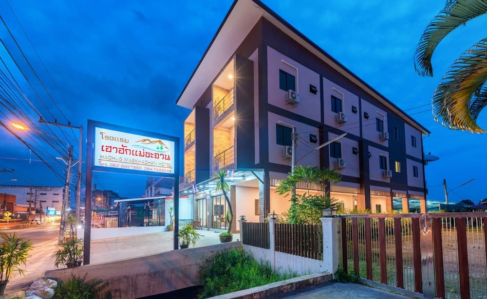 A modern hotel illuminated at dusk features a welcoming entrance, lush greenery, and a prominent signboard, ideal for travelers seeking accommodation in a tropical urban setting.