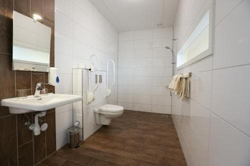 A clean, accessible hotel bathroom with white tiled walls, wooden flooring, a sink, toilet, shower area, and neatly hung towels, ideal for travelers seeking comfort and convenience.