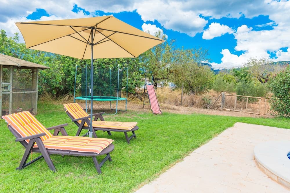 A serene backyard oasis features striped lounge chairs under a beige umbrella, a trampoline, and slide, set against lush greenery and a picturesque mountain backdrop, ideal for family vacations or relaxing getaways.