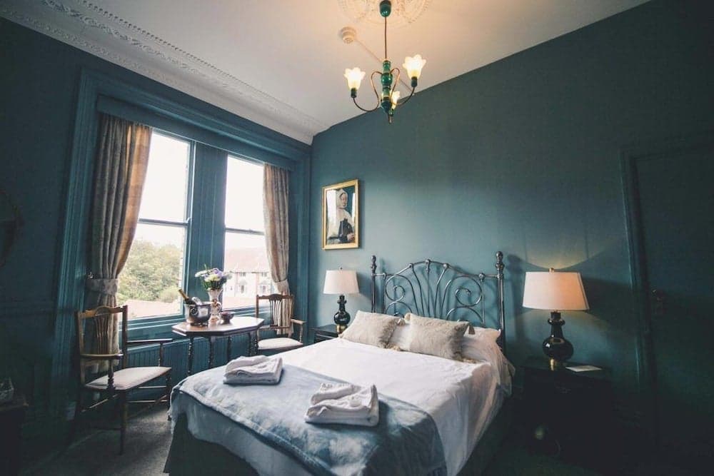 A cozy, vintage-inspired hotel room features teal walls, a wrought iron bed, elegant lamps, and a bright window view, ideal for travelers seeking a charming and relaxing stay.