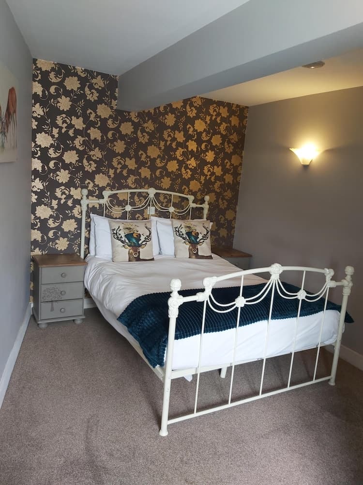 A cozy bedroom with a white iron bed adorned with stag-patterned pillows, set against a floral wallpaper backdrop, exudes warmth under soft lighting, ideal for travelers seeking a charming and comfortable retreat.