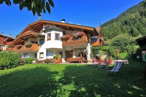 A charming alpine chalet with balconies adorned by flowers sits amidst lush greenery under a clear blue sky, offering a serene retreat ideal for nature lovers seeking relaxation.