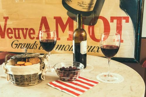 A cozy bistro table set with red wine glasses, a bottle of champagne, bread, olives, and a vintage "Veuve AMTOT" sign evokes a romantic Parisian dining experience perfect for travelers seeking authentic French charm.