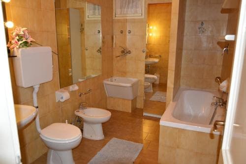 A cozy, warmly lit bathroom with beige tiles, a bathtub, bidet, toilet, and decorative mirror, ideal for travelers seeking comfort and cleanliness in their accommodations.
