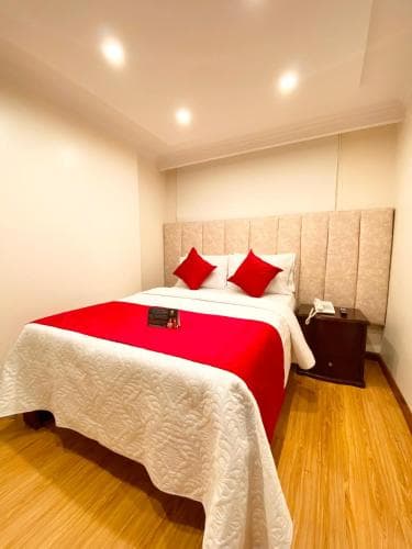 A cozy hotel room features a neatly made bed with red accents, warm wooden flooring, and soft lighting, ideal for travelers seeking comfort and relaxation.
