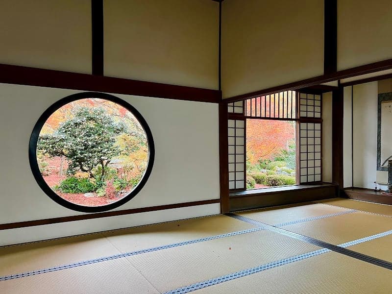 A serene tatami room with a circular window framing autumn foliage offers a tranquil Japanese tea ceremony experience in Kyoto's historic setting.