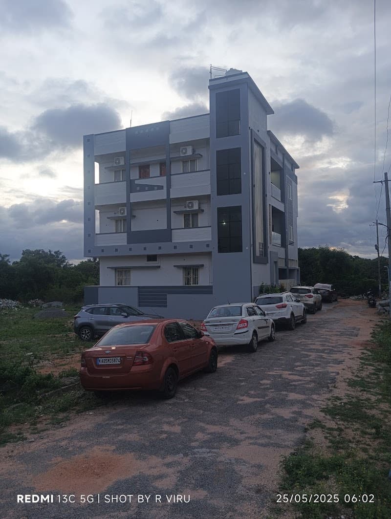 A modern multi-story building stands under a cloudy sky in a serene rural setting near Hampi, Karnataka, with parked cars lining the gravel path, ideal for travelers seeking accommodation close to historical sites.
