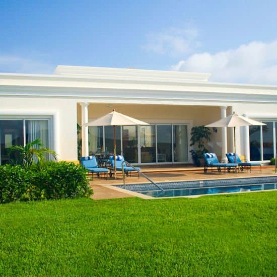 A serene luxury villa with a pristine pool, lush green lawn, and shaded lounge areas under umbrellas invites travelers seeking a tranquil tropical retreat.