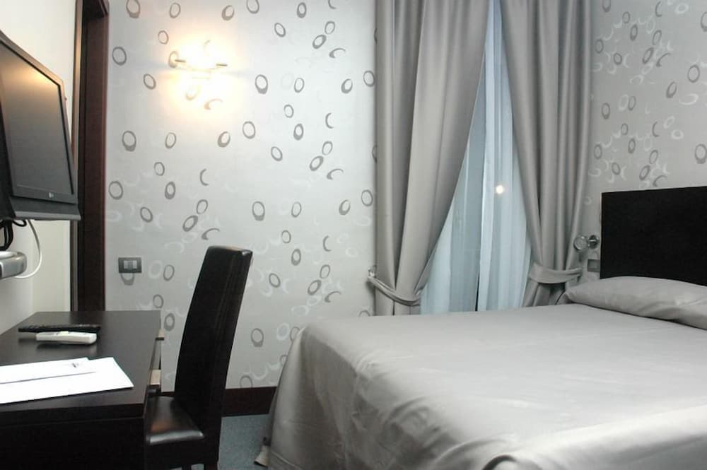A cozy hotel room features a neatly made bed, patterned wallpaper, a desk with a chair, and soft lighting, ideal for travelers seeking comfort and functionality.