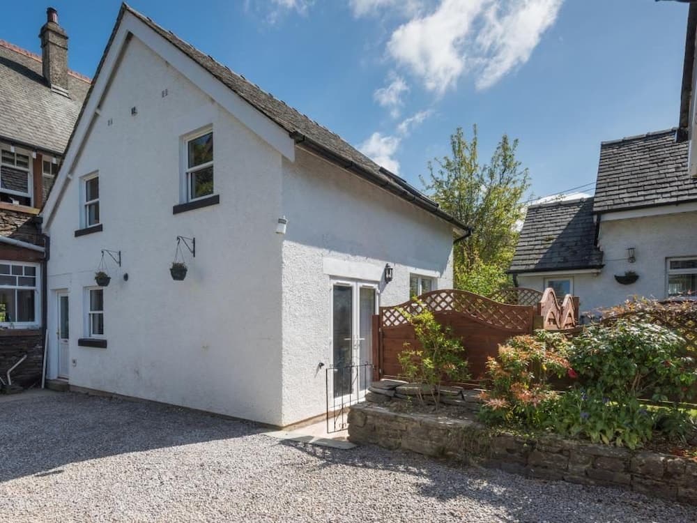 A charming white cottage with a gravel driveway, wooden gate, and lush garden invites travelers to enjoy a peaceful countryside retreat under a bright blue sky.