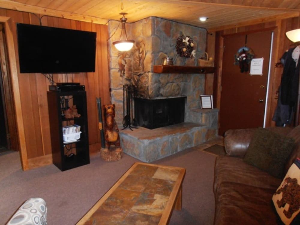 A cozy cabin living room features a stone fireplace, wooden walls, a leather couch, and a TV, creating a warm and inviting atmosphere ideal for a relaxing getaway.