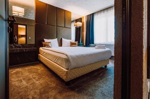 A cozy hotel room features a neatly made bed with plush pillows, warm lighting, and a large window draped in curtains, creating an inviting atmosphere ideal for travelers seeking comfort and relaxation.