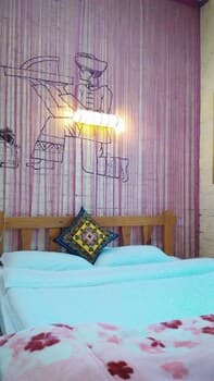 A cozy bedroom with a wooden bed adorned in blue bedding and a floral-patterned blanket, set against a pink striped wall featuring a whimsical black-and-white mural, evokes a serene and artistic atmosphere ideal for travelers seeking a unique and tranquil accommodation experience.