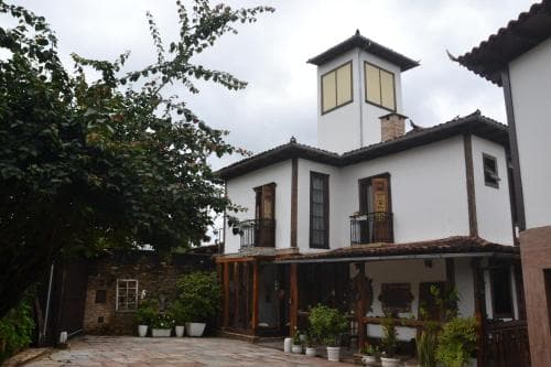 A charming white house with wooden balconies and lush greenery exudes a cozy, rustic atmosphere, perfect for travelers seeking a tranquil retreat in a quaint village setting.