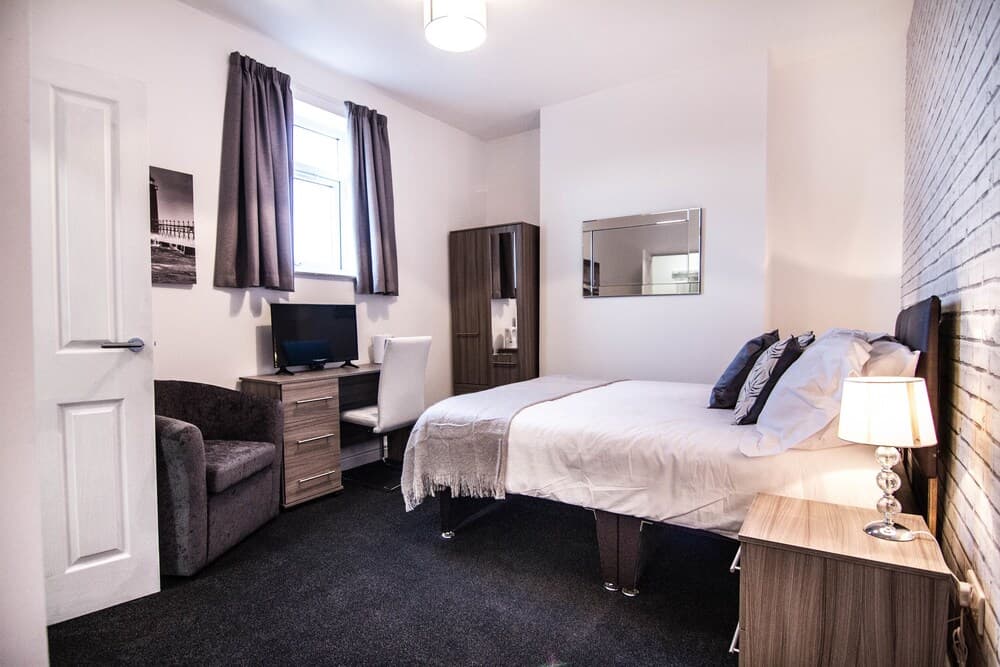 A cozy hotel room features a neatly made bed beside a desk with a TV, a plush armchair, and a brick accent wall, creating a comfortable and modern atmosphere ideal for travelers seeking relaxation.