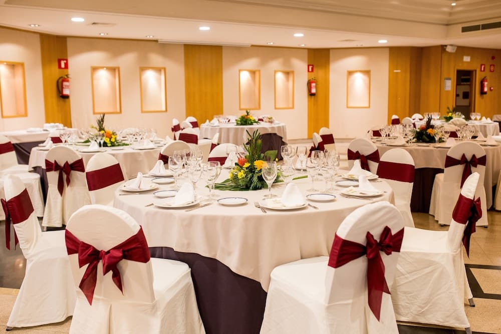A spacious banquet hall adorned with elegant white tables, chairs tied with red bows, and vibrant floral centerpieces creates an inviting atmosphere perfect for wedding receptions or formal gatherings.