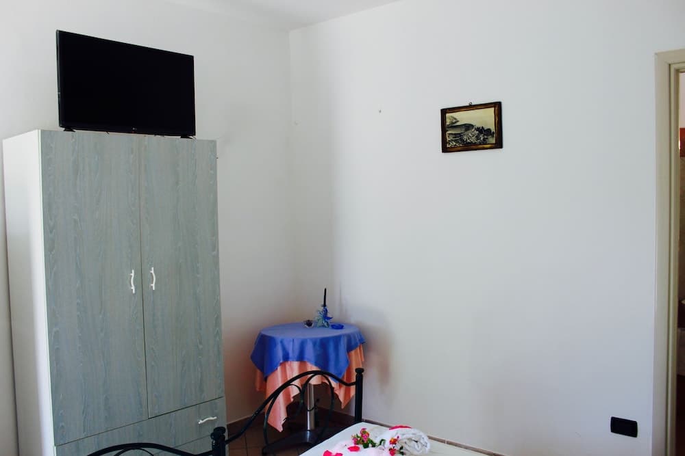 A cozy hotel room features a wooden wardrobe topped with a TV, a small table draped in colorful cloths, and a neatly made bed adorned with flowers, ideal for travelers seeking comfort and simplicity.