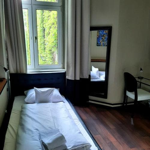 A cozy hotel room with a neatly made bed, hardwood floors, a large window overlooking greenery, and a stylish mirror reflects the serene ambiance ideal for travelers seeking comfort and relaxation.