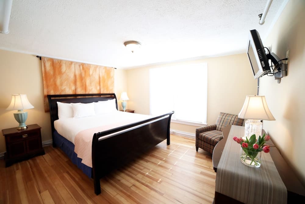 A cozy hotel room with warm lighting, wooden floors, a sleek black bed, plaid armchair, and vibrant tulips on the dresser creates an inviting atmosphere ideal for travelers seeking comfort and relaxation.