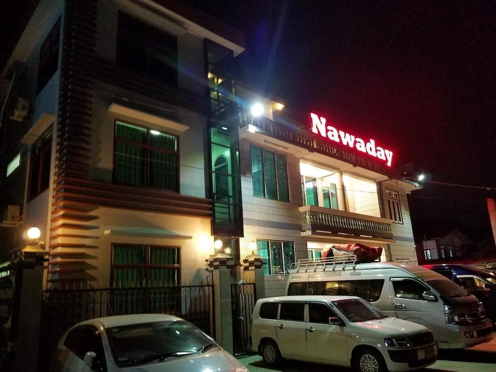 A vibrant night scene showcases a modern building illuminated by bright lights and a glowing "Nawaday" sign, surrounded by parked cars, ideal for travelers seeking accommodation or nightlife in an urban setting.