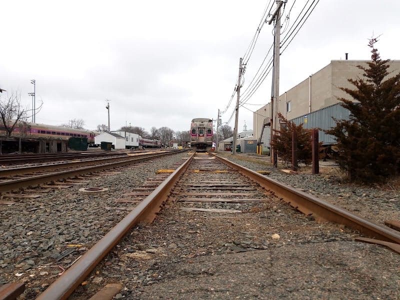 A serene railway scene in Rockport, MA, features multiple tracks leading towards an approaching train under overcast skies, surrounded by industrial buildings and utility poles, ideal for travelers exploring the coastal town's transportation history.